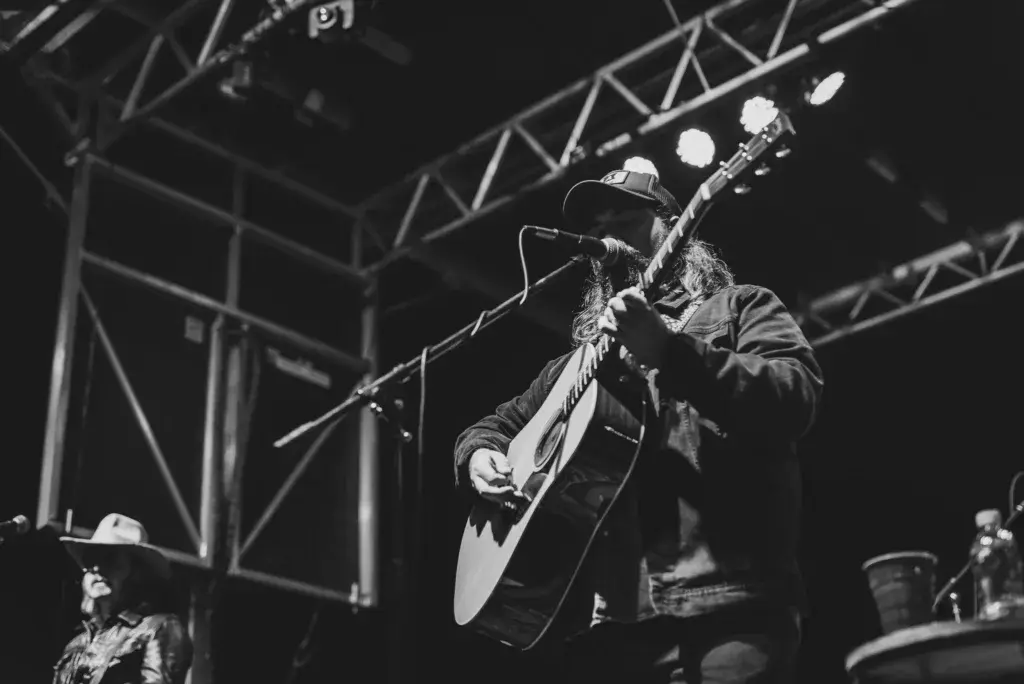 A performer wearing a cowboy hat plays an acoustic guitar on stage under bright lights.