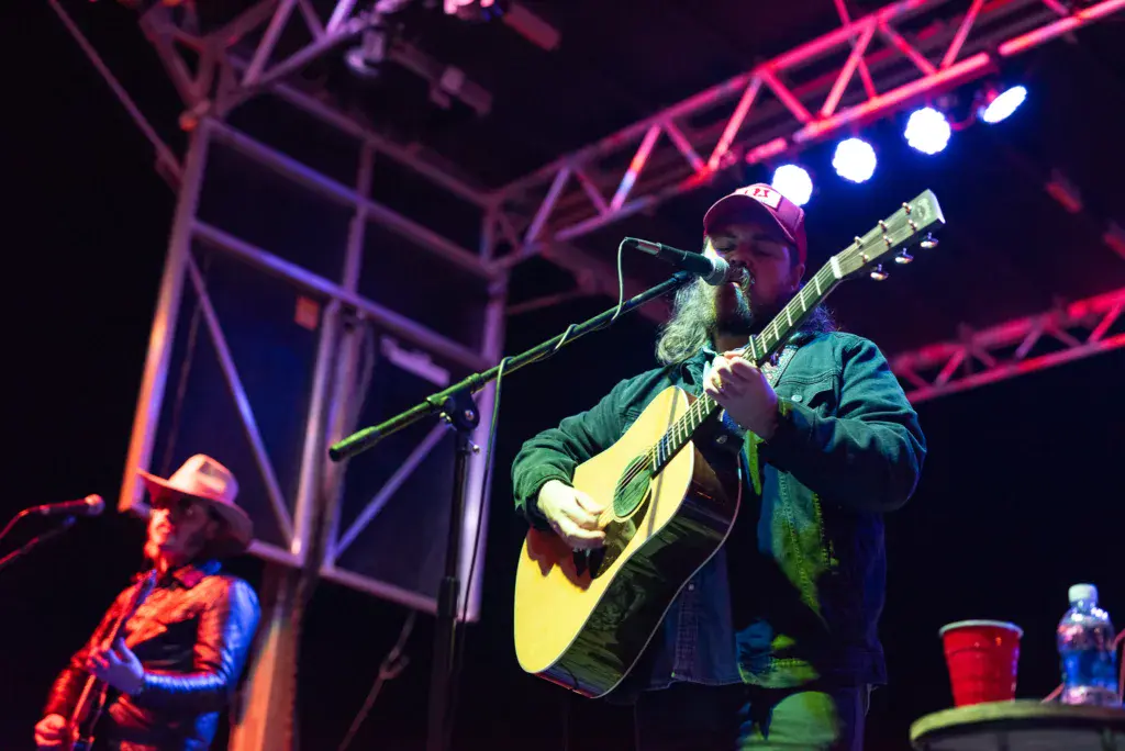 Bearded guitarist wearing a cap performs on stage with acoustic guitar under colorful stage lighting.