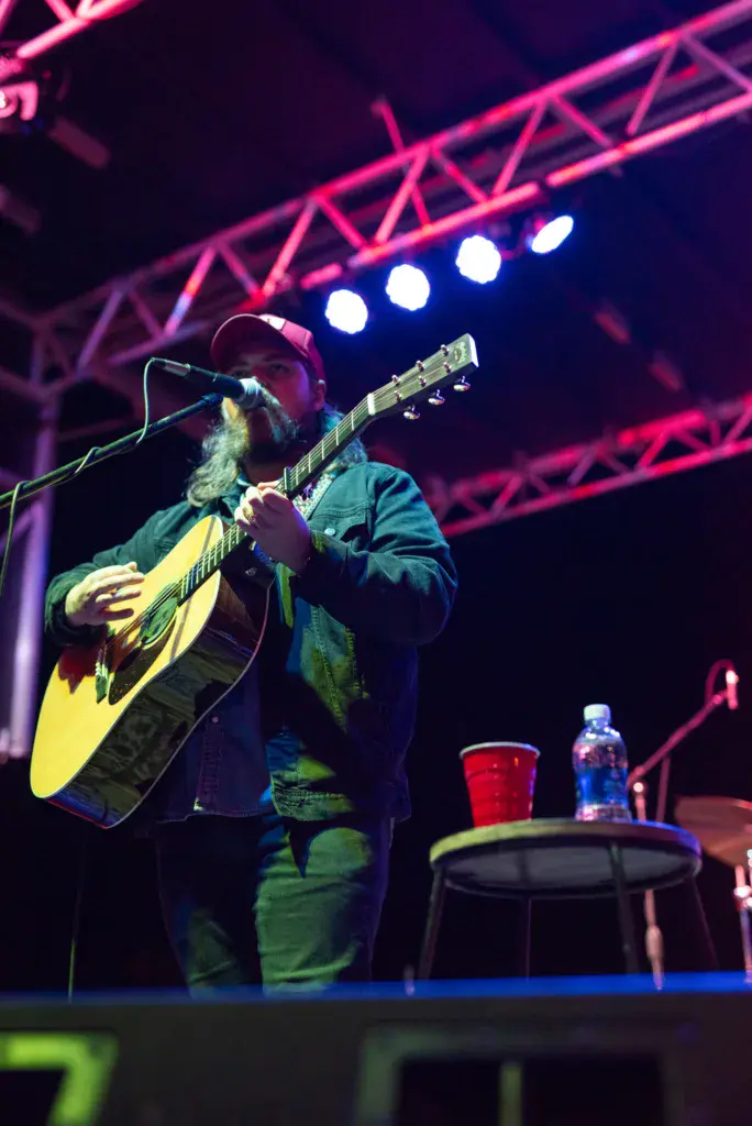 A bearded performer plays an acoustic guitar at the Dancing Rabbit Music Festival stage under pink truss lighting.