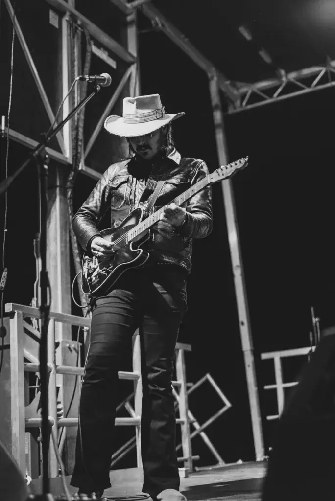 A guitarist wearing a white cowboy hat performs on stage under bright lights during an evening concert.