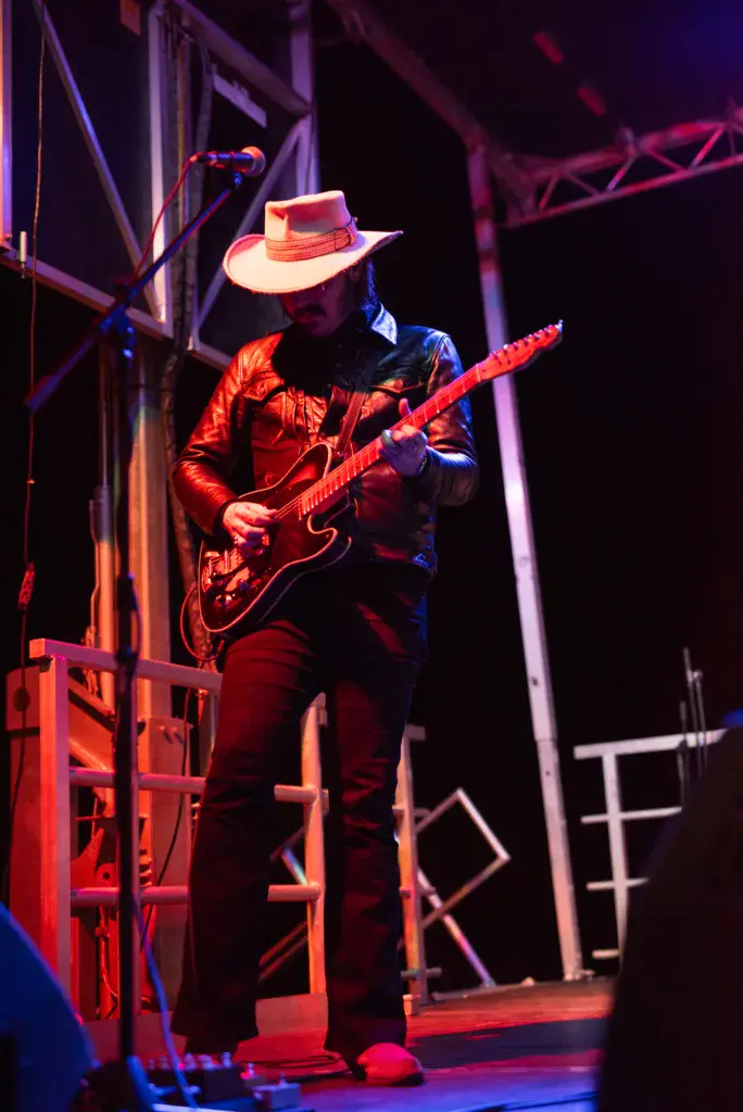 A guitarist in a red shirt and cowboy hat performs on stage under purple and red lighting.