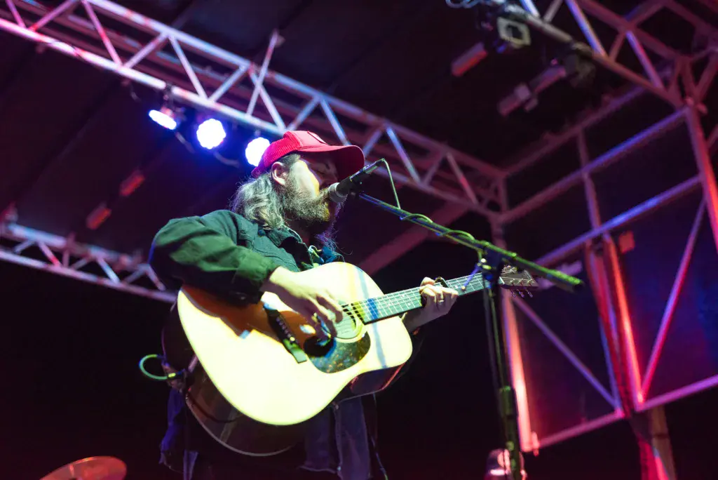 A musician with a beard wearing a red cap performs with an acoustic guitar on stage under colorful purple and blue lighting.