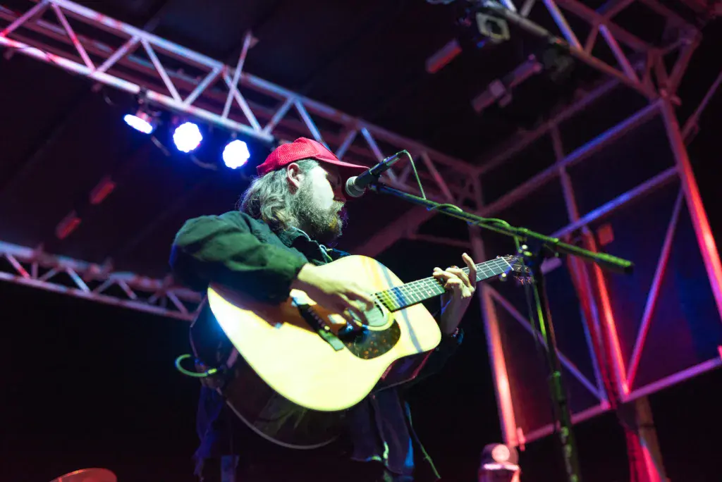A bearded musician wearing a red cap plays an acoustic guitar and sings into a microphone on an illuminated stage.