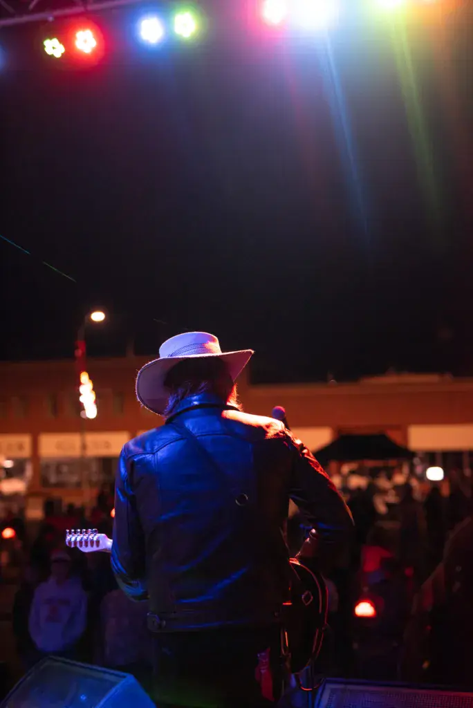A performer wearing a cowboy hat plays guitar on stage under colorful lighting at night.