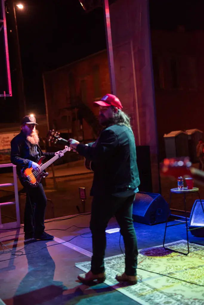 Two musicians perform on a stage with colorful pink and blue lighting at an evening concert.