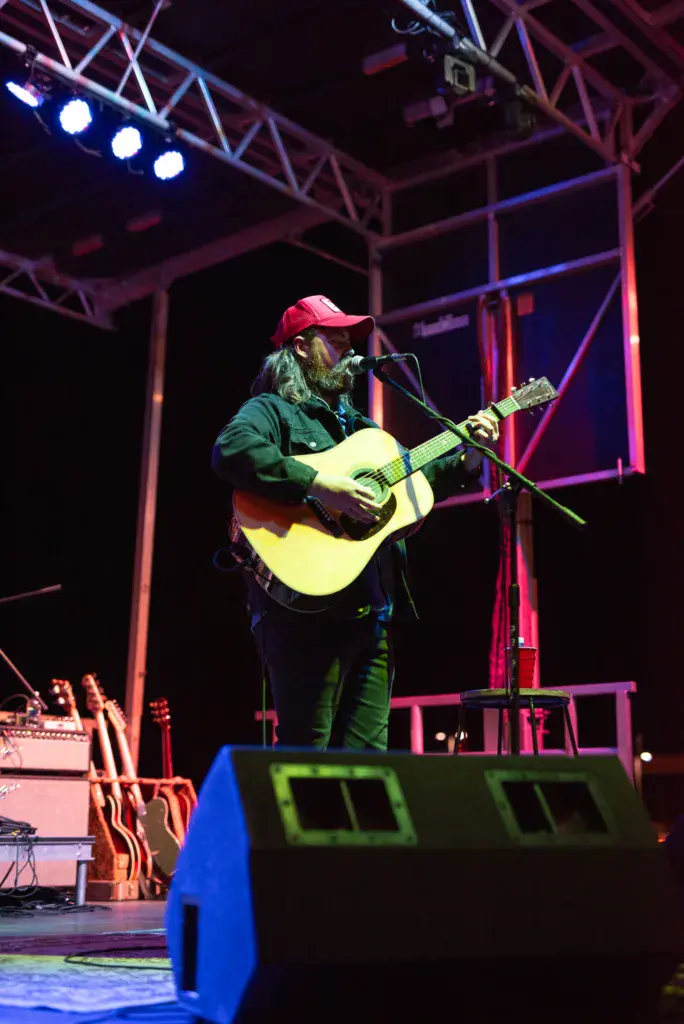 A solo performer plays acoustic guitar on an illuminated stage at night.