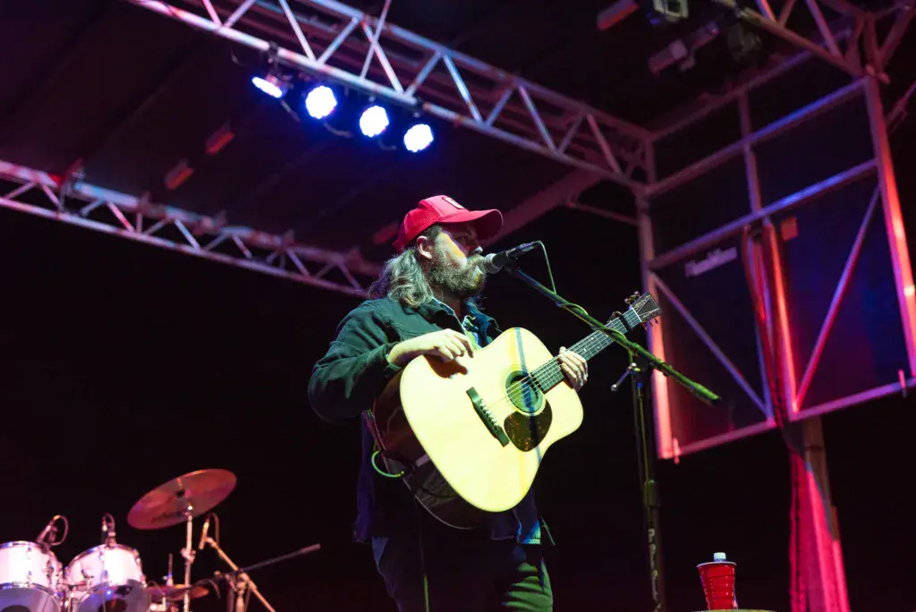 Guitarist in red cap performs with acoustic guitar on stage under colorful lighting.