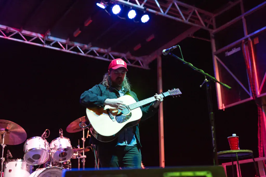 A performer plays acoustic guitar on stage under blue and red lighting at an outdoor music festival.