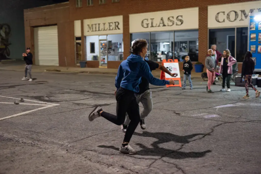 A person in a blue jacket runs through a parking lot while holding an orange sign, with brick buildings and spectators visible in the background.