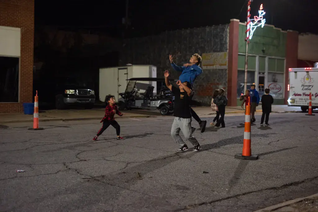 A person in a blue shirt tosses a child in the air during an evening outdoor event in a parking lot.