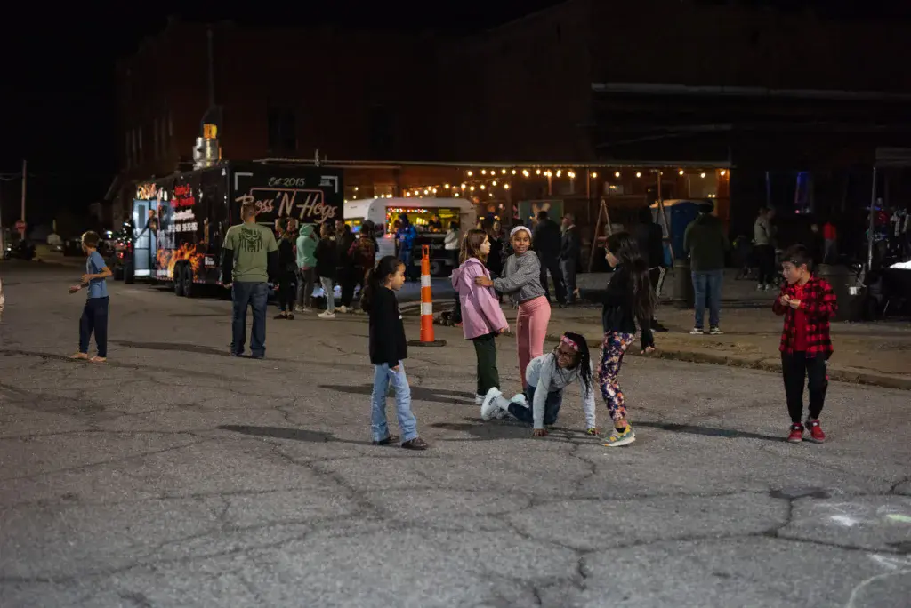 Festival attendees gather in a parking lot at night near illuminated vendor booths and signage.