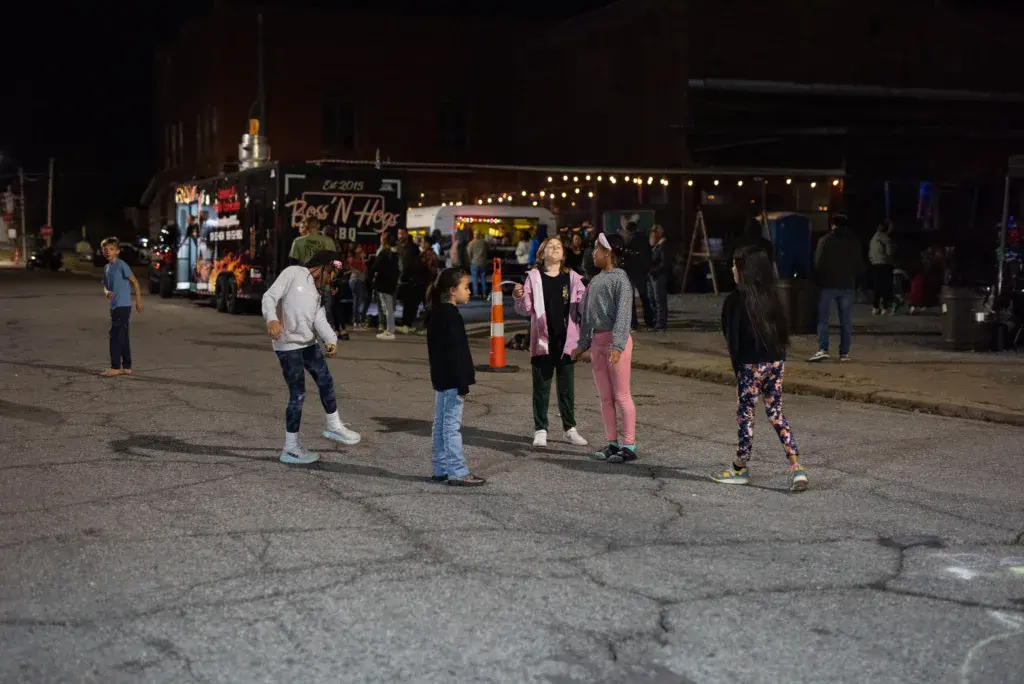 Evening festival crowd gathered in parking area near illuminated food vendor booth labeled Boss N Hoss BBQ.