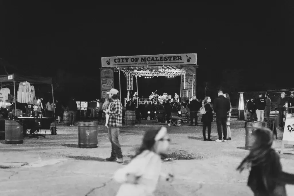 Black and white photograph of the City of McAlester stage at night with festival attendees and vendors scattered across the grounds.