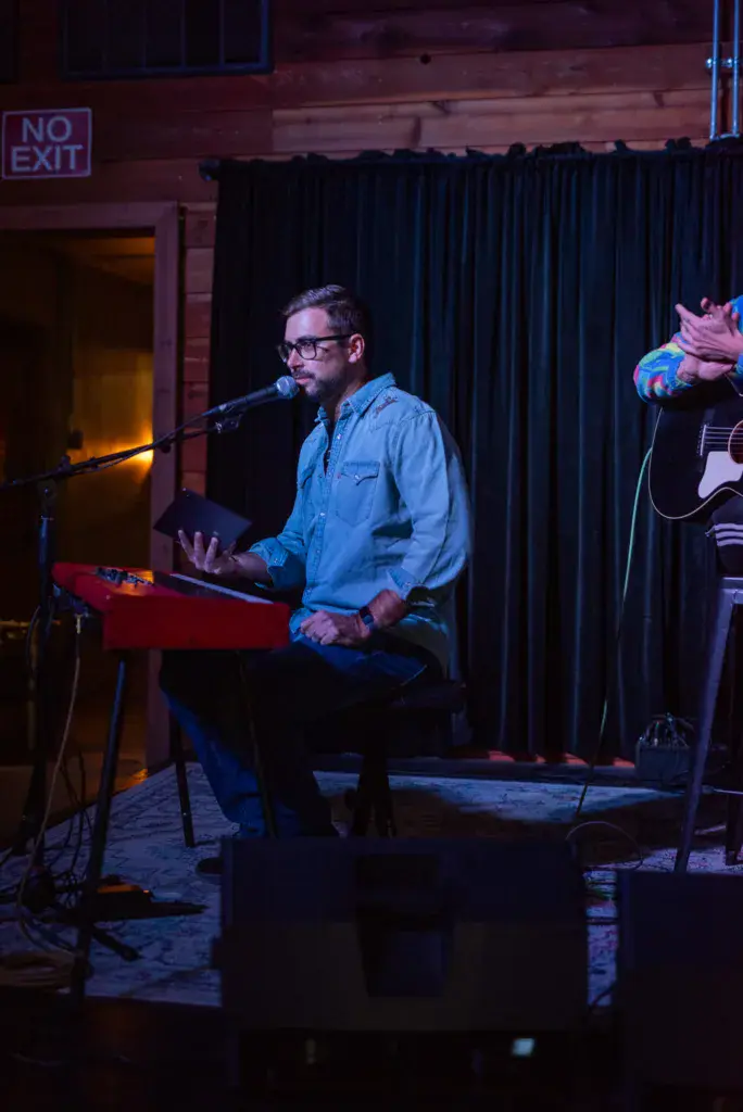 A musician wearing glasses and a light blue shirt plays keyboard and sings into a microphone on stage.