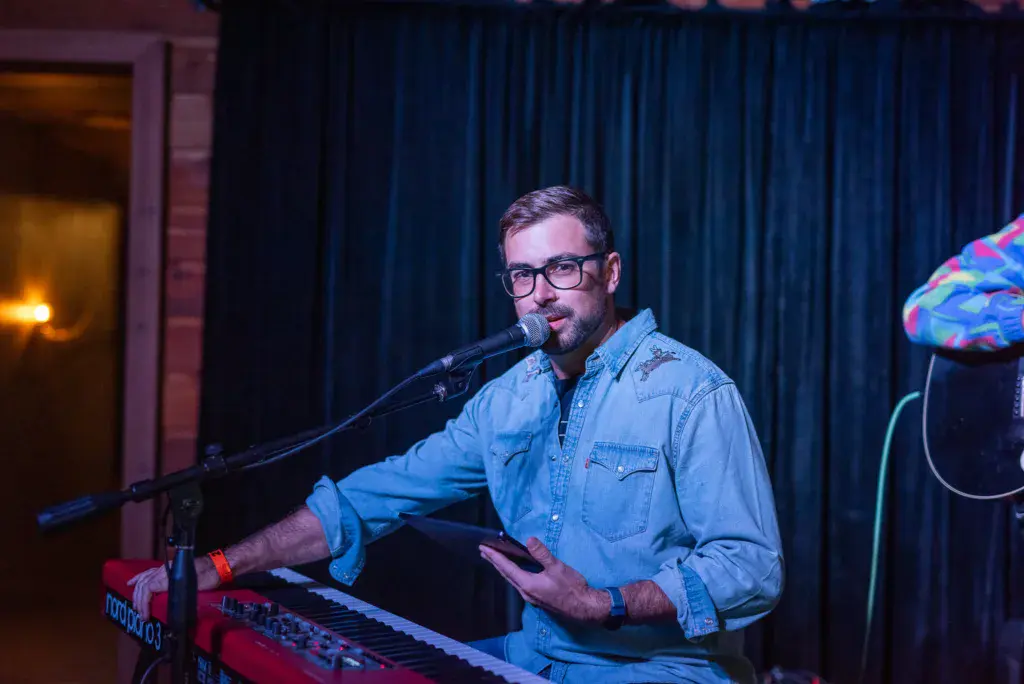 A performer wearing glasses and a denim shirt plays keyboard and sings into a microphone on stage.