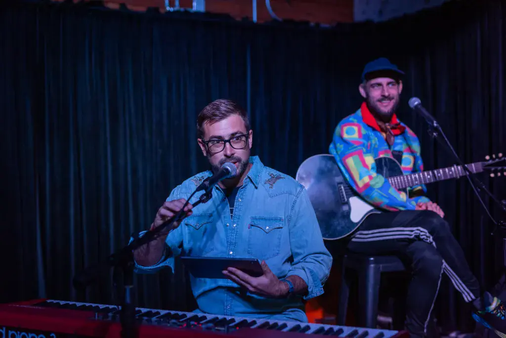 Two musicians perform on stage at an indoor venue with blue curtains behind them.