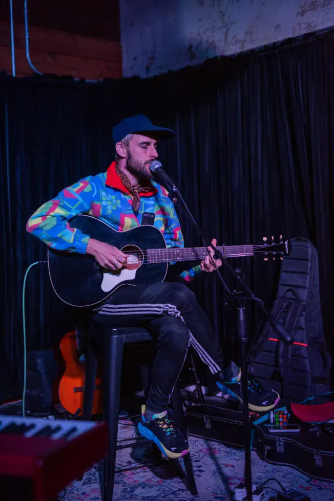 A bearded musician in a colorful patterned jacket sits on stage playing an acoustic guitar into a microphone.