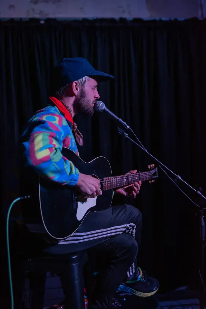 A bearded musician wearing a colorful jacket and blue cap plays acoustic guitar and sings into a microphone on stage.