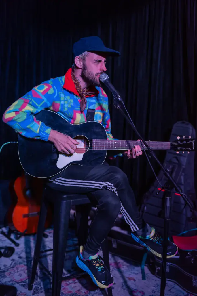 A bearded musician in a colorful geometric jacket performs with an acoustic guitar at an indoor venue.
