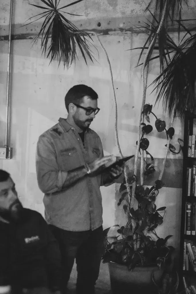 A man wearing sunglasses and a collared shirt holds a tablet while standing near hanging plants indoors.