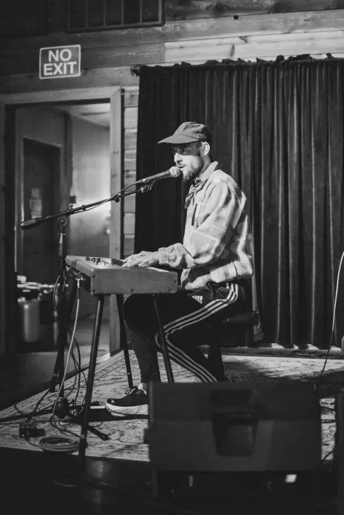 A performer with a beard and cap sings into a microphone while playing a keyboard instrument on an indoor stage.