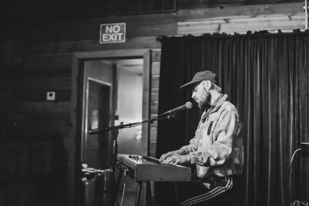 Musician performing at a microphone while playing a keyboard at an indoor venue with a no exit sign visible in the background.