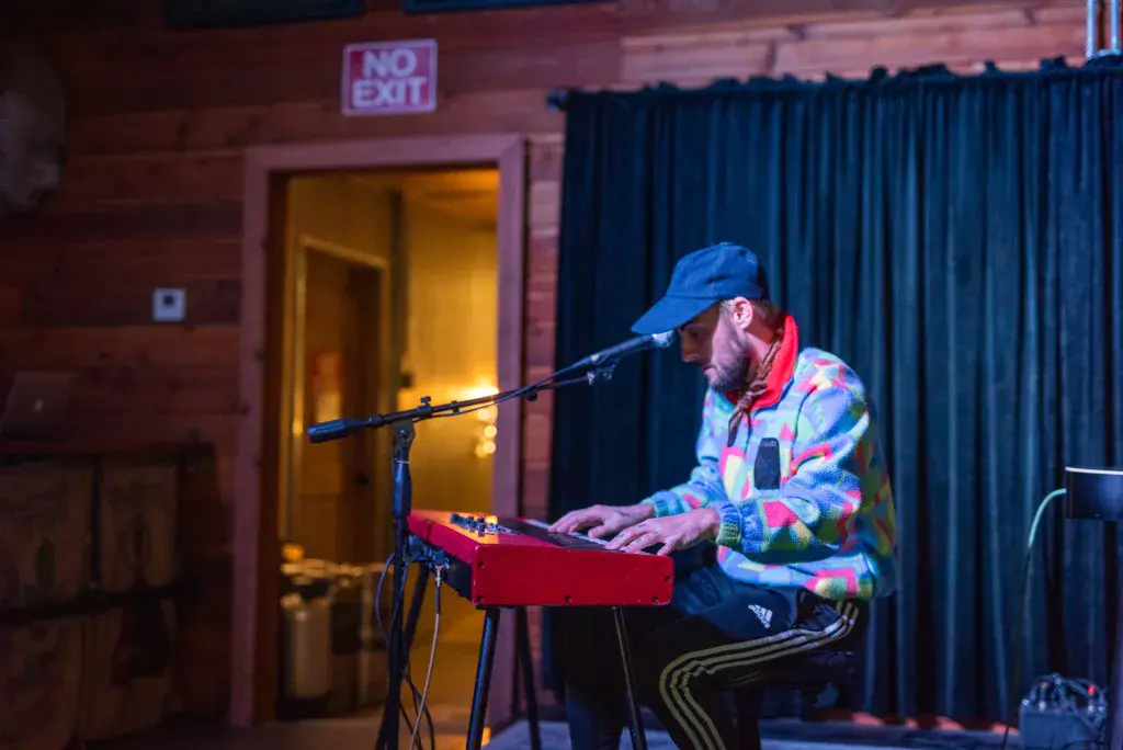 A performer in a colorful patterned jacket and blue cap plays a red keyboard at an indoor stage venue.