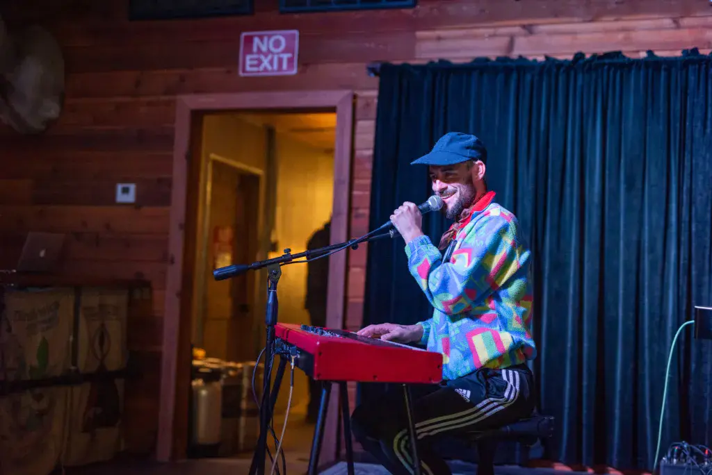 A performer sings into a microphone while playing a red keyboard on an indoor stage.