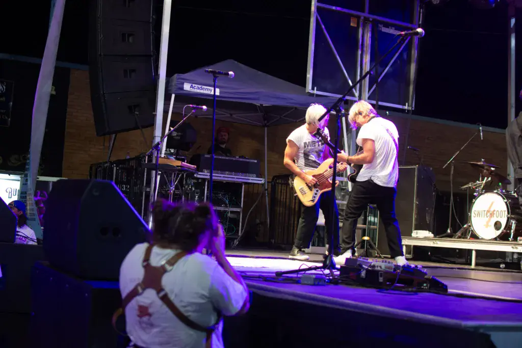 Band performs on stage at night with purple lighting while crowd members watch from the foreground.