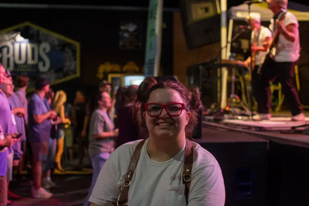 A smiling festival attendee in glasses stands in front of a lit stage with musicians and crowd in the background.