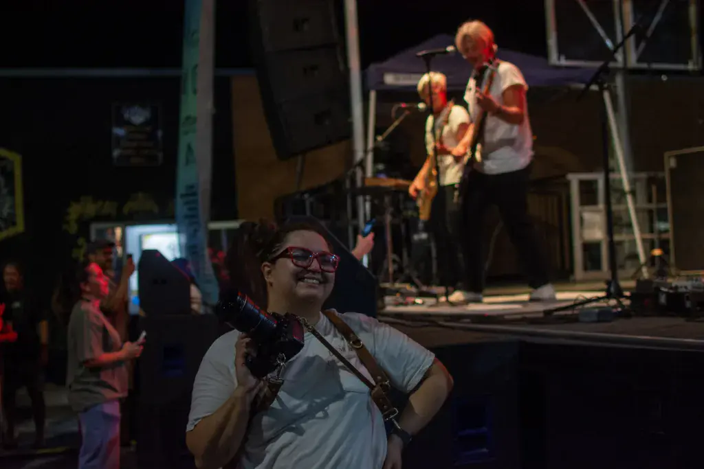 A festivalgoer in glasses smiles while watching a band perform on an illuminated stage at night.