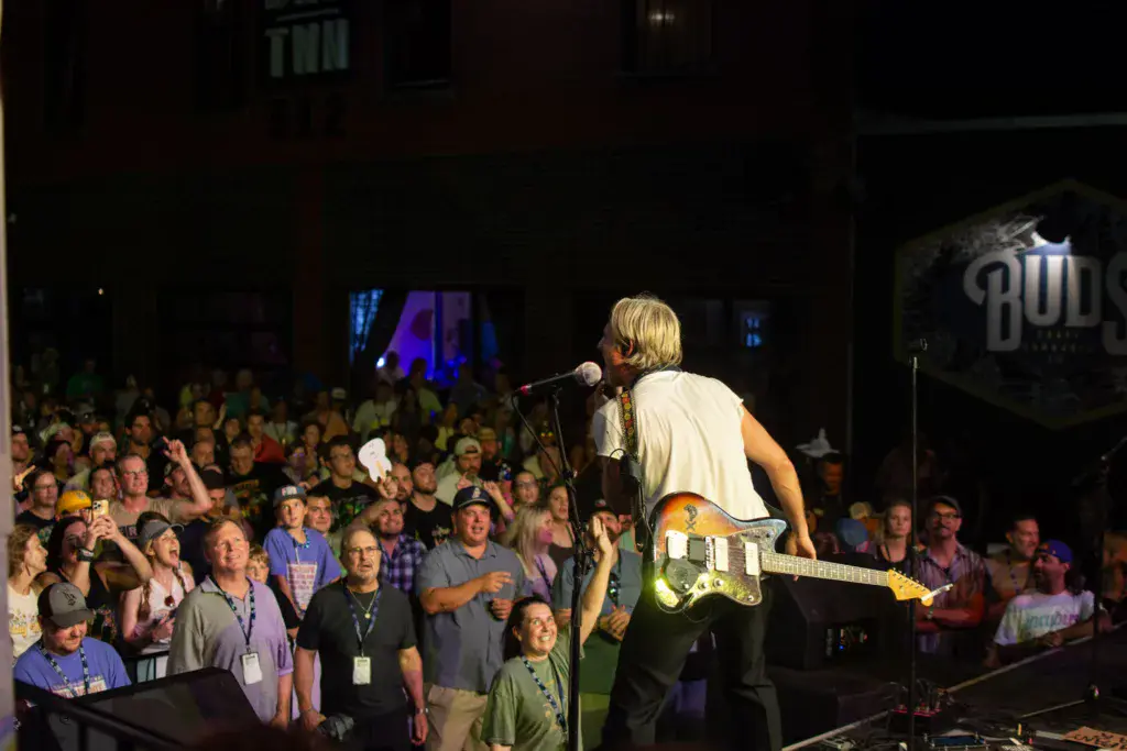 A musician with blonde hair performs on stage with an electric guitar while a large crowd watches from the audience.