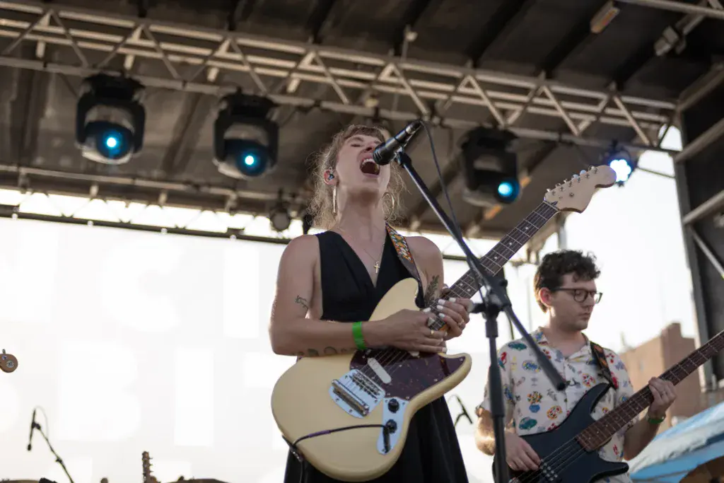 A guitarist and bassist perform on stage under blue stage lights at an outdoor music festival.