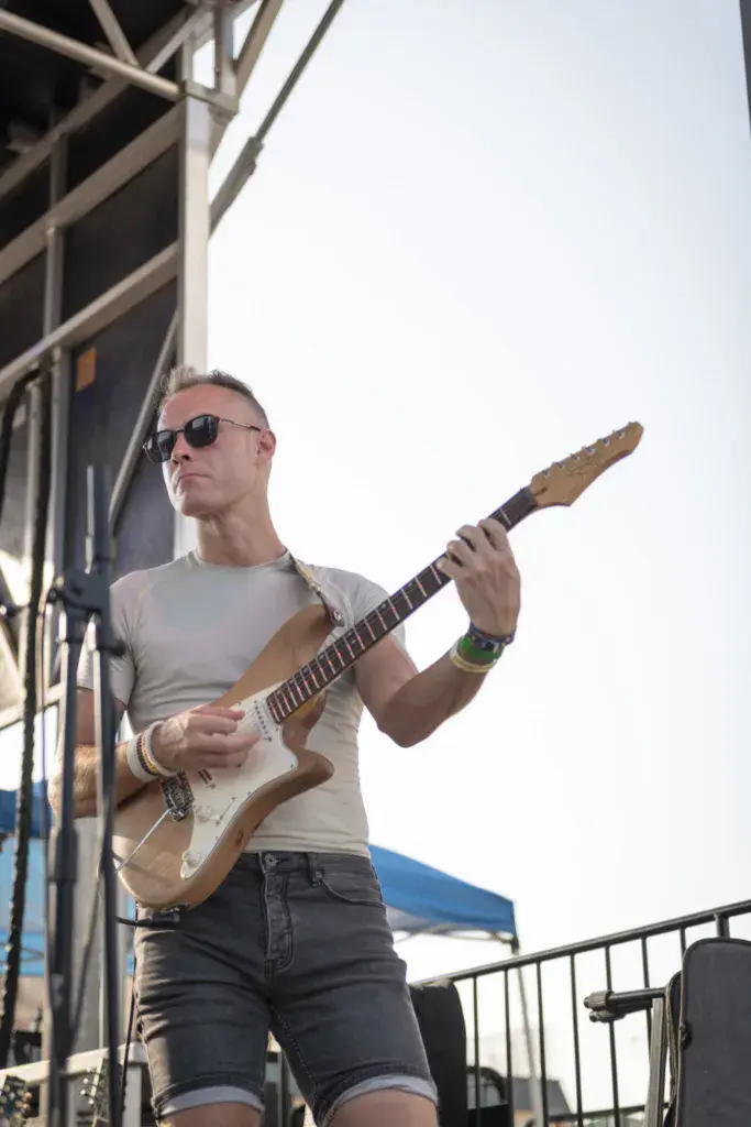 A guitarist wearing sunglasses performs on stage with a light-colored electric guitar.