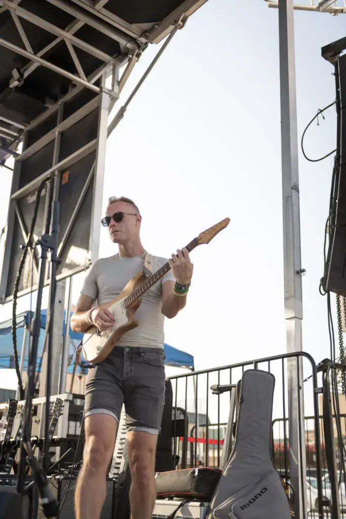 Guitarist wearing sunglasses and light gray shirt performs on outdoor festival stage under clear sky.