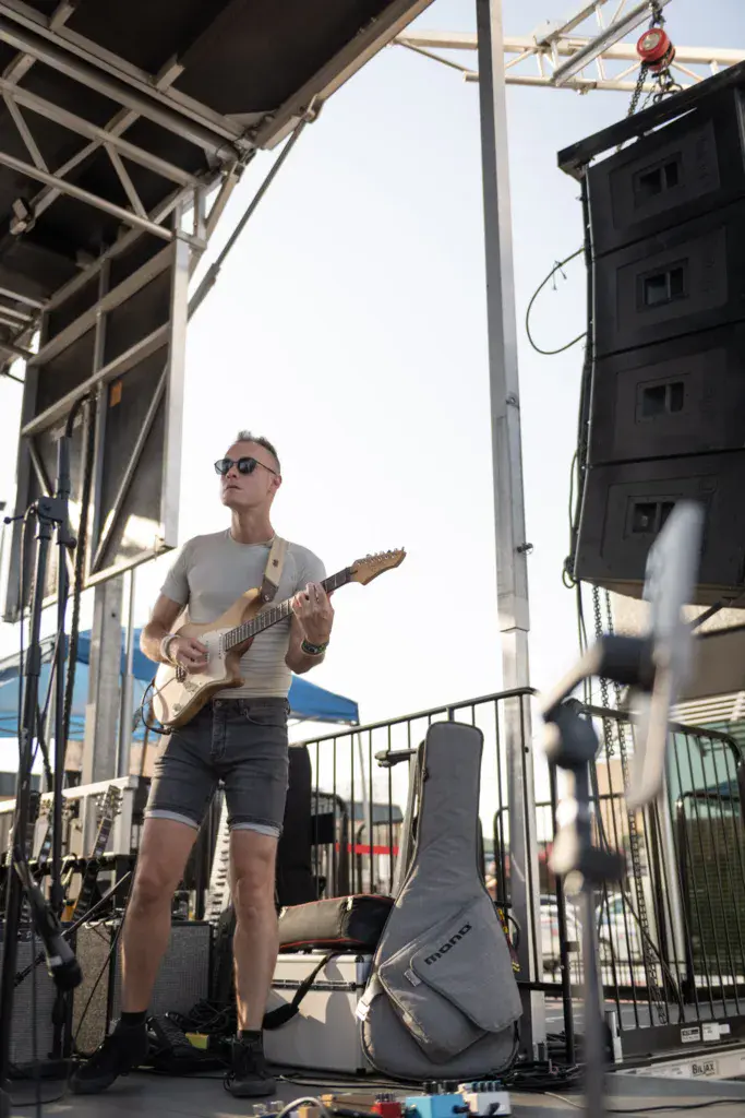 Guitarist performing on stage at an outdoor music festival under a metal structure with amplifier and equipment nearby.