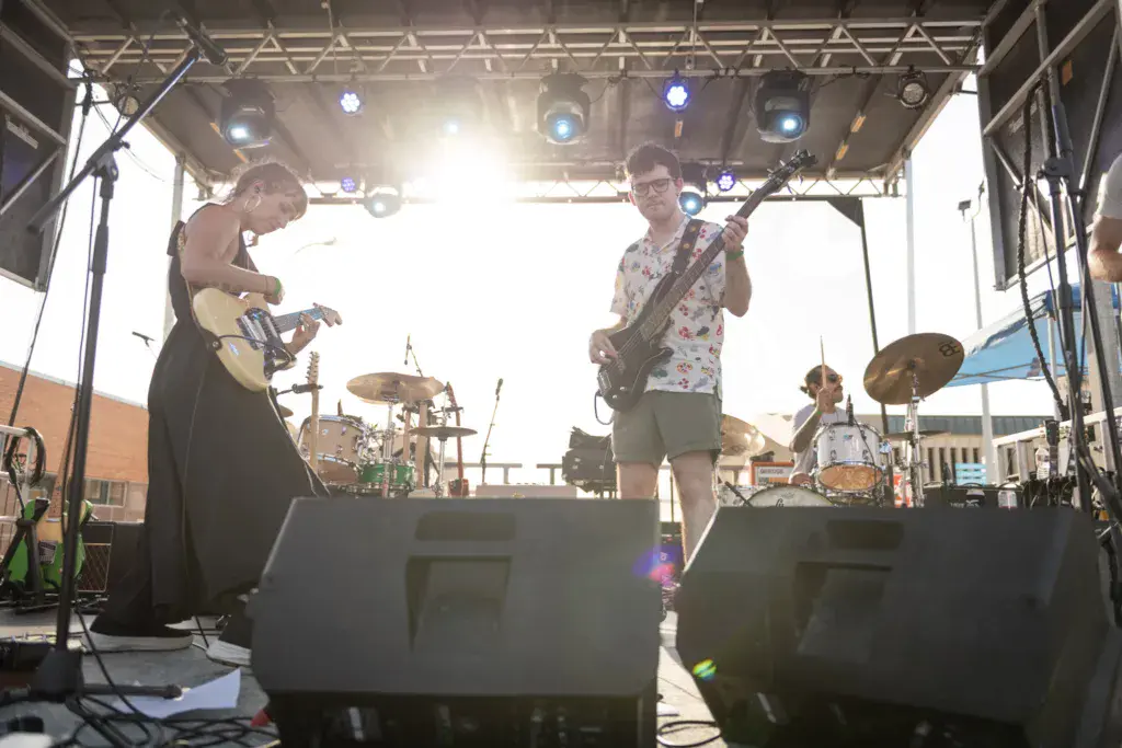 A band performs on an outdoor stage with guitars and drums under bright stage lights.