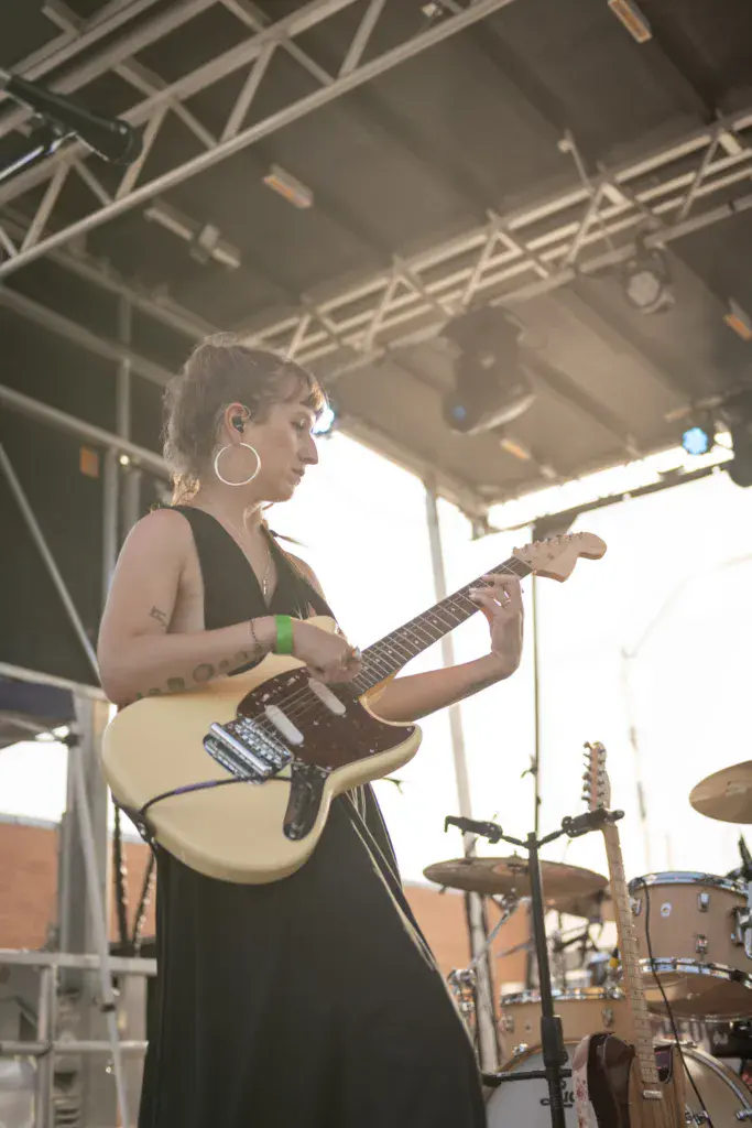 A musician performing on stage with an electric guitar under a metal truss structure at an outdoor festival.