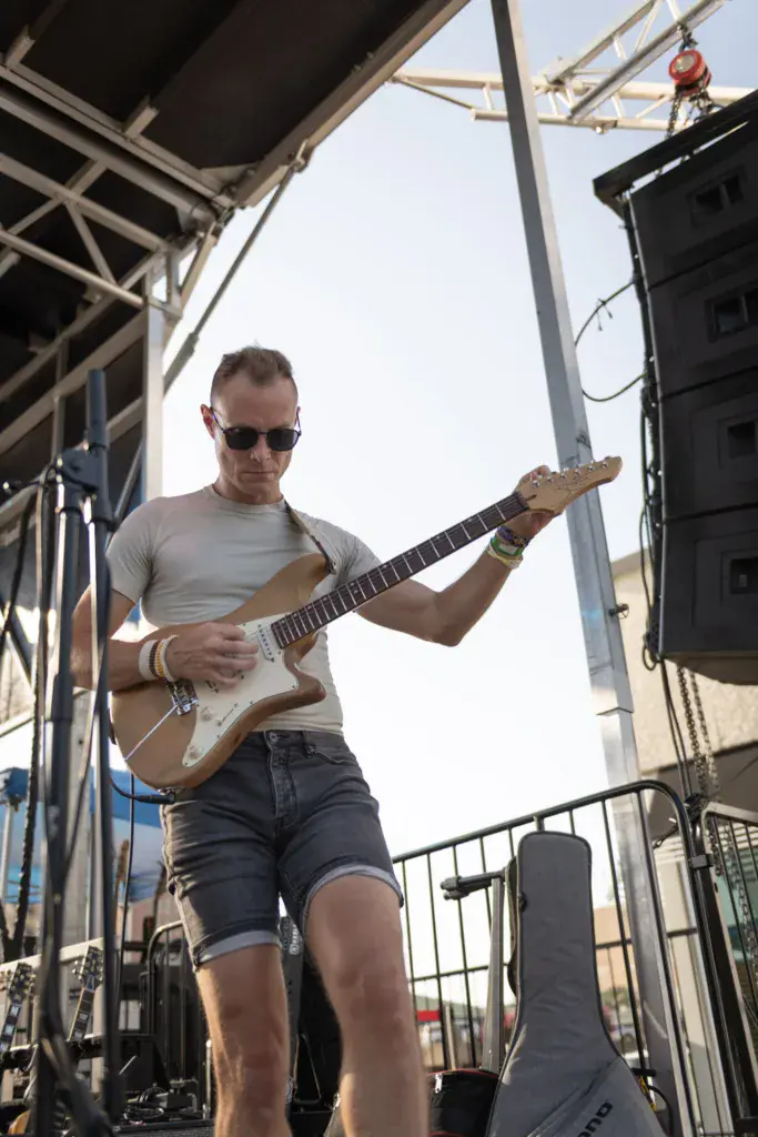 Guitarist performing on stage under a metal framework structure during daytime.