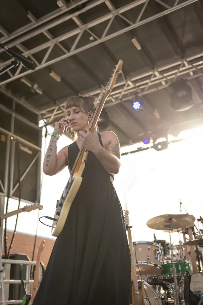 A performer sings into a microphone while playing guitar on stage under industrial truss lighting.