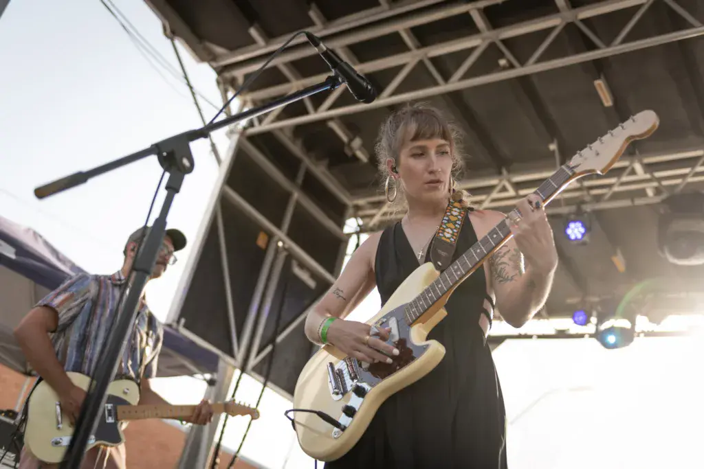 Guitarist performing on stage at an outdoor music festival under a metal truss structure.