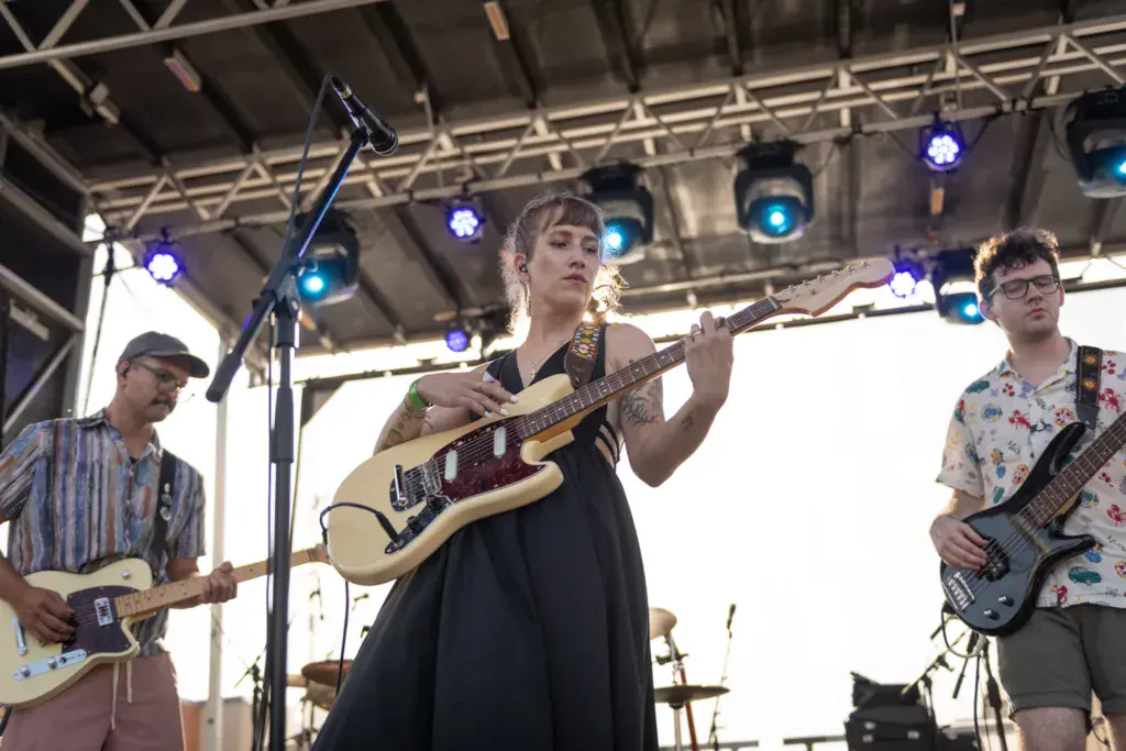 A three-piece band performs on stage under blue lighting at an outdoor music festival.