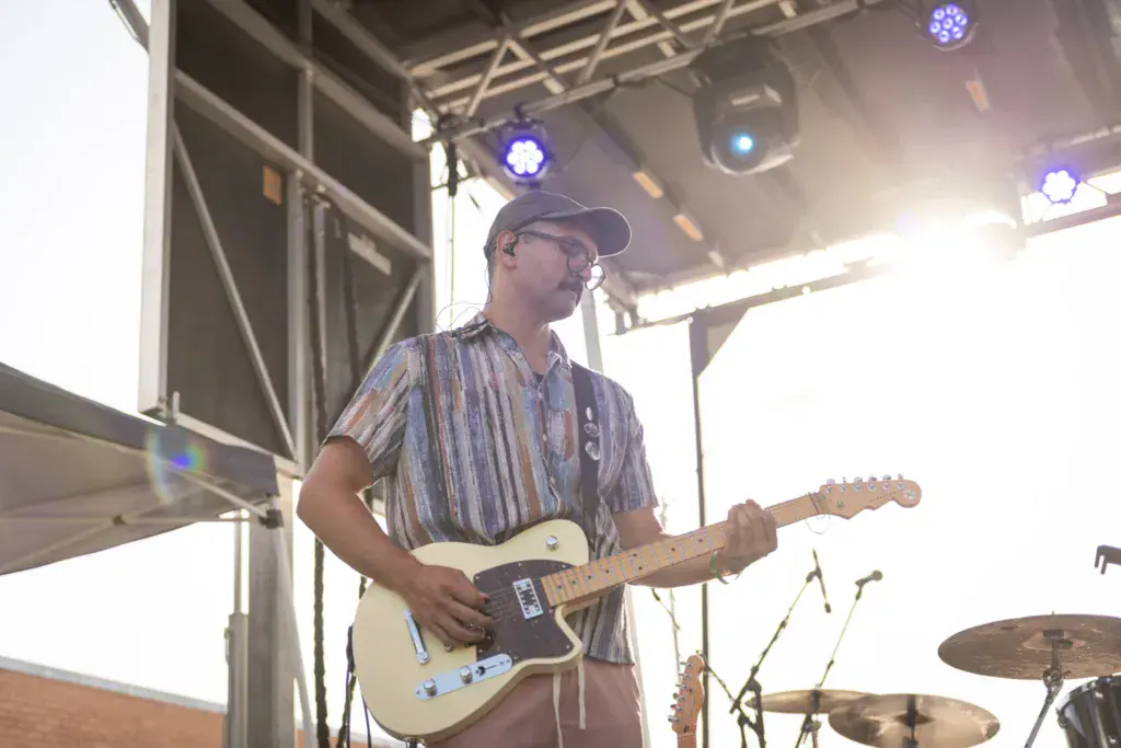 Musician playing electric guitar on stage under blue stage lights at an outdoor festival.
