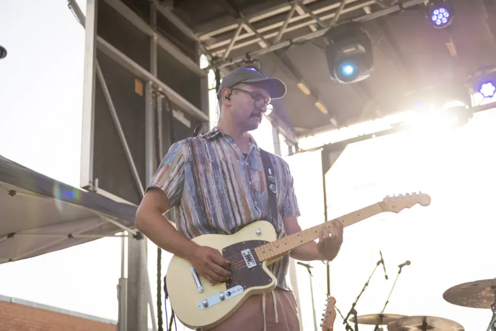 A guitarist in a striped shirt and cap performs on stage under bright lights at an outdoor music festival.