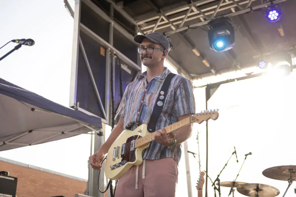 A guitarist performs on stage under bright lights at the festival, wearing a striped shirt and cap.