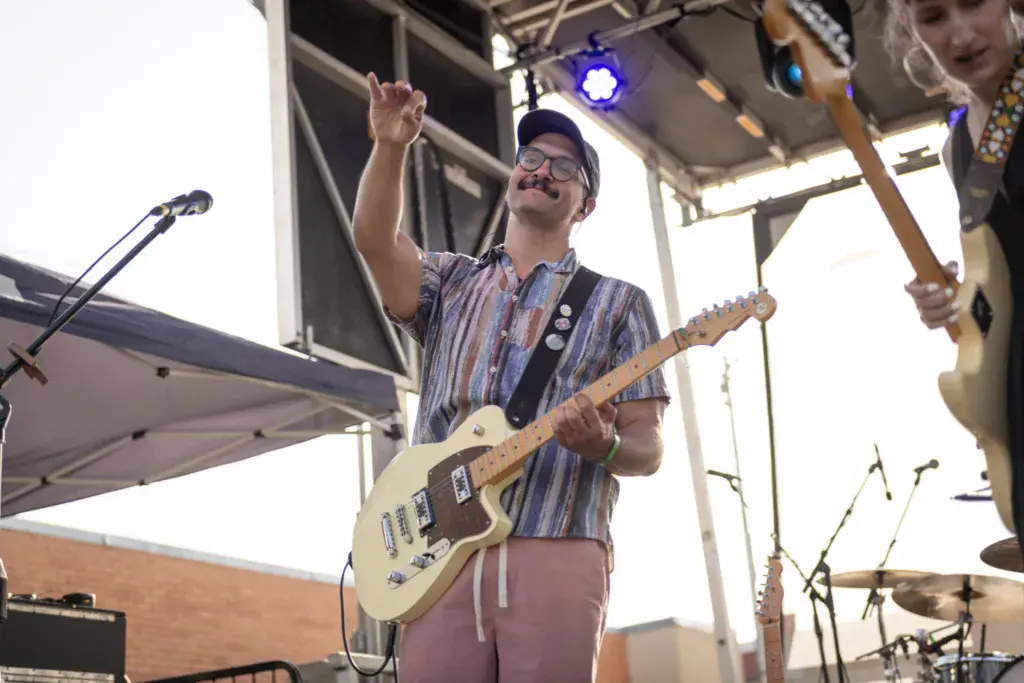 Guitarist with glasses performing on stage under a tent canopy, singing and playing a cream-colored electric guitar.