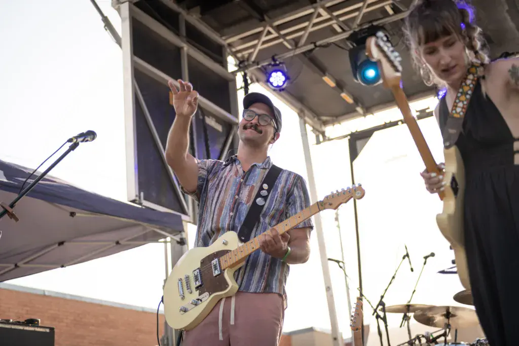 Guitarist with glasses and mustache performing on stage at an outdoor music festival under a metal canopy.