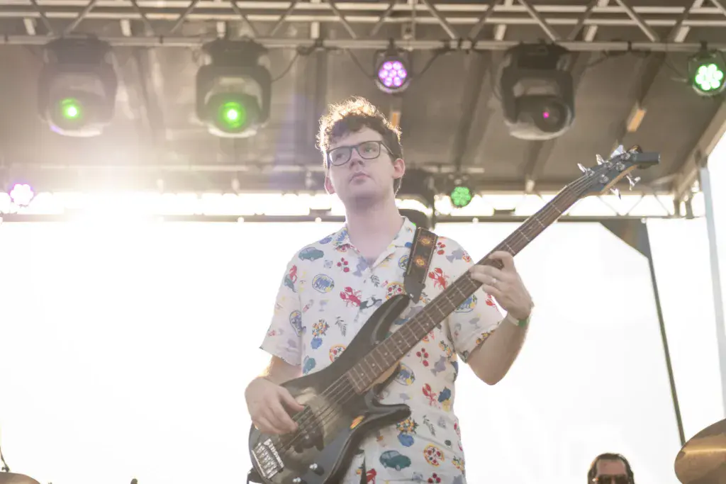 Guitarist wearing floral shirt and glasses performs on stage with colorful stage lights overhead.