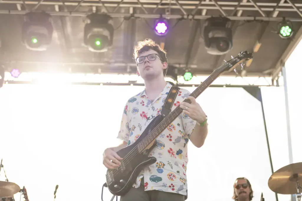 A bass guitarist wearing glasses and a floral shirt performs on stage under colored stage lights.