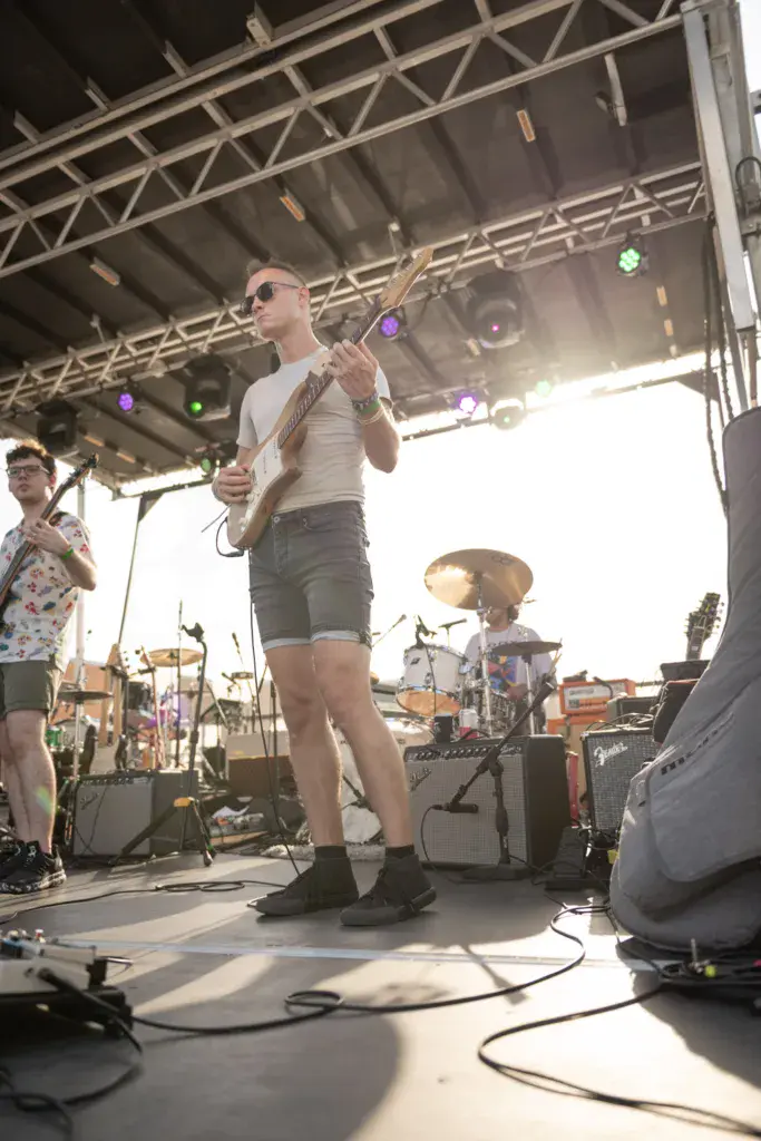 A guitarist performs on stage under a metal truss structure during daytime at an outdoor festival.